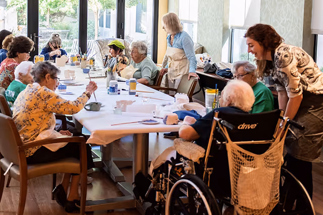A group of elderly individuals seated around a long table engaged in a painting activity, assisted by two caregivers in a bright room with large windows.