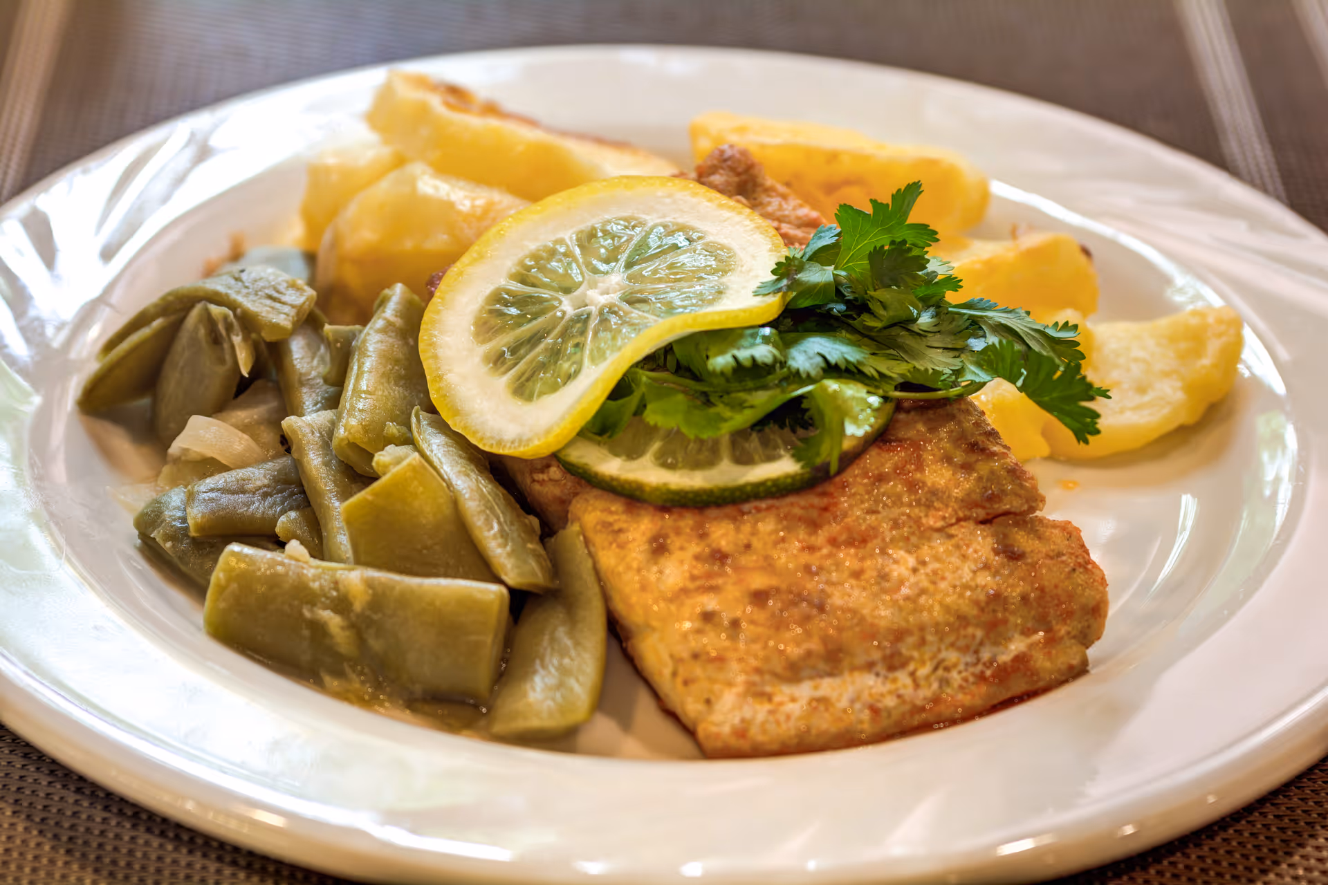 A white plate with a serving of cooked green beans, a piece of seasoned fish topped with lemon slices and fresh parsley, and several pieces of cooked potatoes.