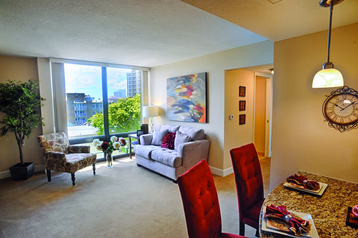 Bright living room with a sofa, patterned armchair, large window showing trees and buildings, and a dining counter with red chairs.