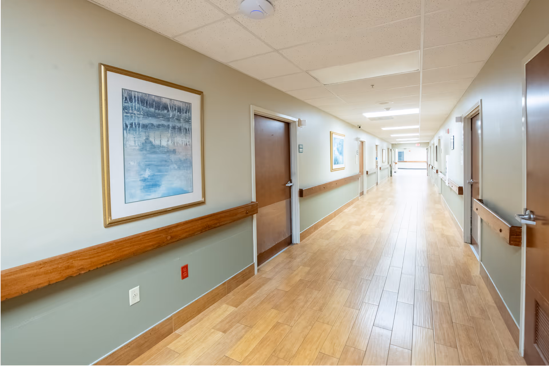 A long, well-lit hallway in a care facility with wooden doors on both sides, handrails along the walls, framed artwork, and light wood flooring.