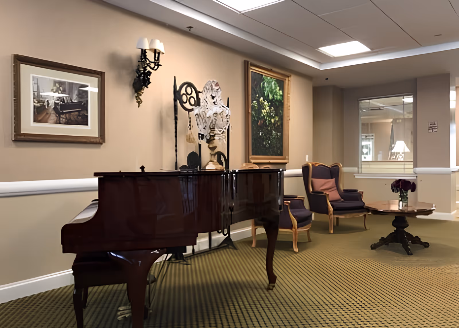 A furnished common room with a glossy grand piano, upholstered chairs, a small table and framed artwork on the walls.