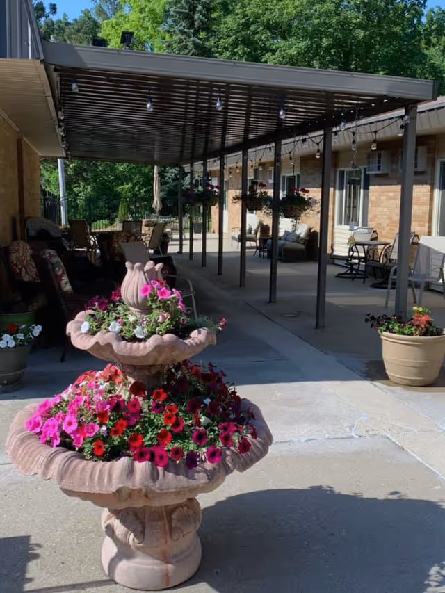 Outdoor patio area at Briarwood Health Care Center featuring a two-tiered stone planter filled with vibrant pink, red, and white flowers in the foreground. The patio has a covered pergola with hanging string lights and several seating arrangements including chairs and tables. The building exterior is visible with brick walls and windows, surrounded by green trees in the background.