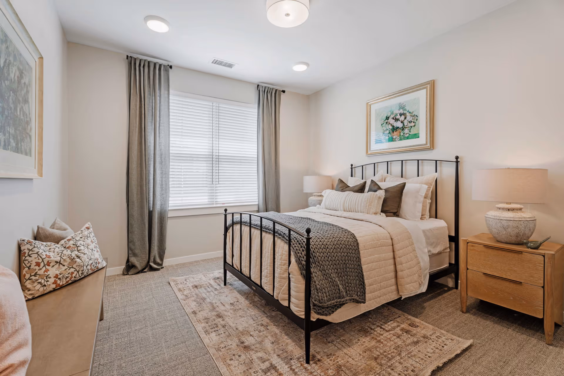 A cozy bedroom with a black metal bed frame, beige bedding, and multiple pillows. There is a wooden nightstand with a lamp on the right side of the bed. A framed floral painting hangs above the bed. The room has a window with closed blinds and gray curtains. A cushioned bench with decorative pillows is placed against the left wall. The floor is carpeted with a patterned area rug under the bed.