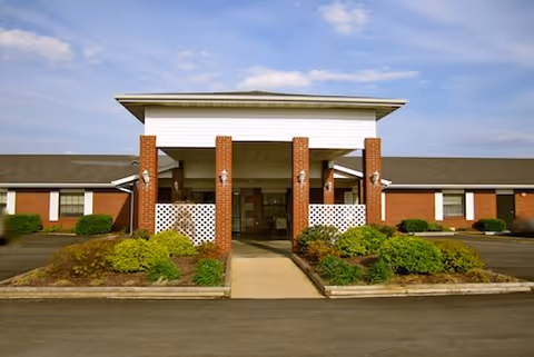 Front entrance of a single-story brick care facility with a covered porte-cochère, shrubs, and a driveway.