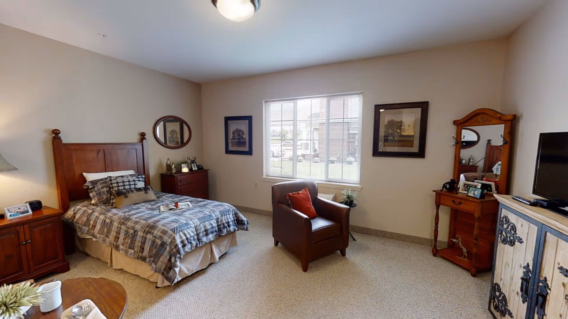 A furnished bedroom with a wooden bed, plaid bedding, an armchair by a window, and wooden dressers and a TV.