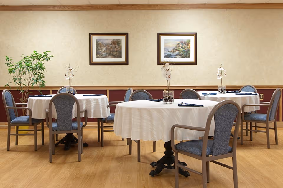 A dining room with round tables covered in white tablecloths, each set with blue napkins and a vase with decorative flowers. The room has wooden flooring, beige walls with a maroon wainscot, two framed landscape paintings on the wall, and a potted plant in the corner.