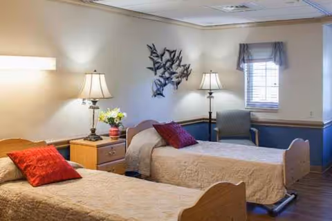 Two twin beds in a cozy resident room with nightstand, lamps, and a chair.