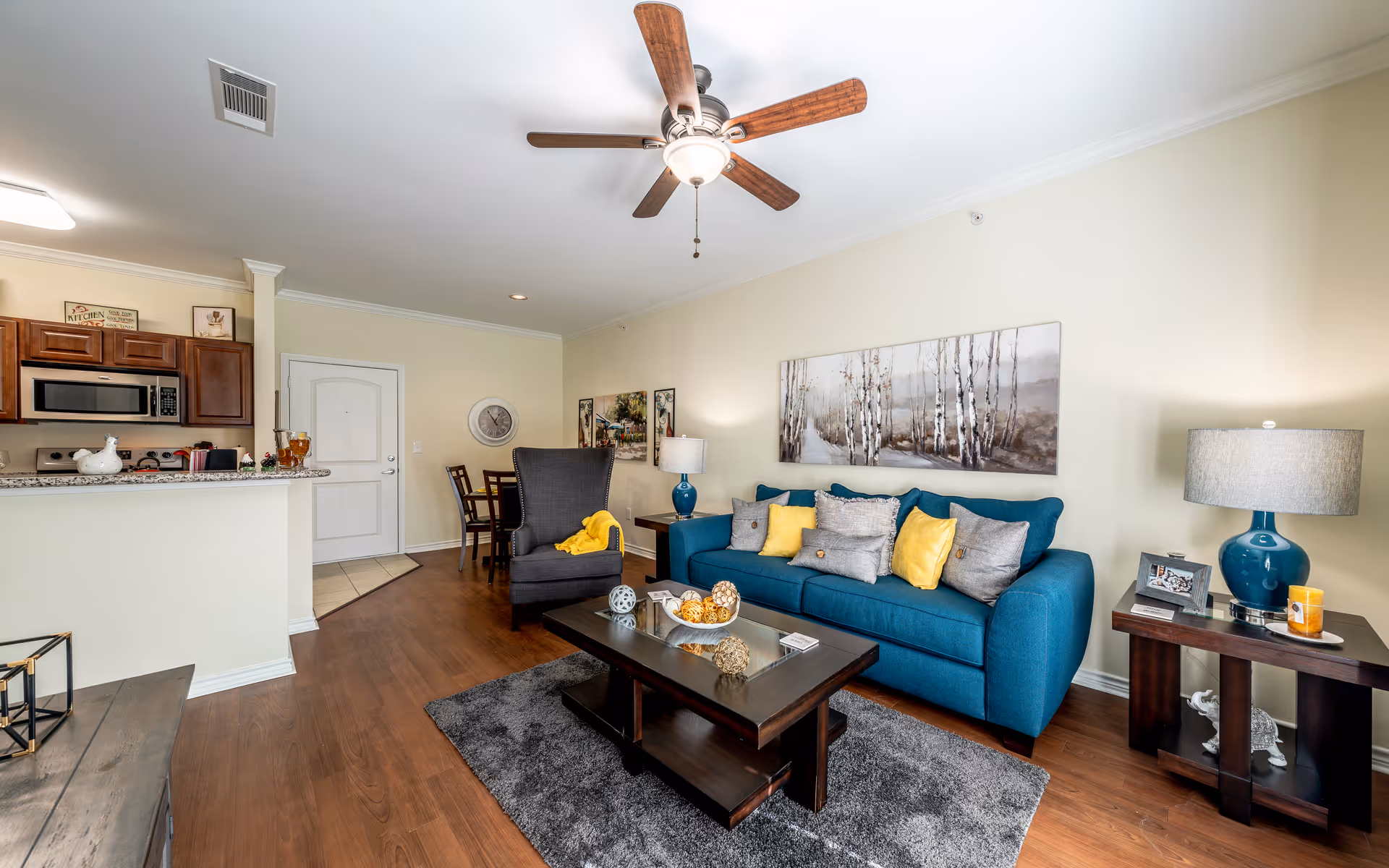 A bright living room with a blue sofa adorned with yellow and gray pillows, a dark wooden coffee table with decorative items, a gray armchair with a yellow throw, and two side tables with blue lamps. The room has wooden flooring, a ceiling fan with lights, and a large painting of birch trees on the wall. In the background, there is a small dining area and a kitchen with wooden cabinets and a microwave.