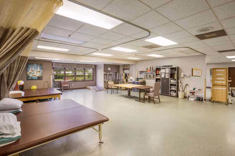 A spacious physical therapy or rehabilitation room with padded treatment tables, chairs, exercise equipment, and storage cabinets. Large windows provide natural light, and the room has a clean, organized appearance with a neutral color scheme.