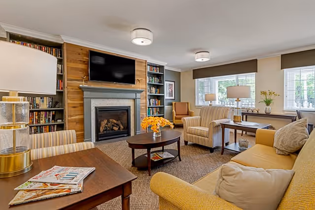 A cozy living room in Brookdale Auburn featuring a fireplace with a mounted TV above it, built-in bookshelves on either side, a round coffee table with a vase of yellow flowers, and comfortable seating including a yellow sofa, striped armchairs, and side tables with lamps. Large windows with blinds allow natural light to fill the room.