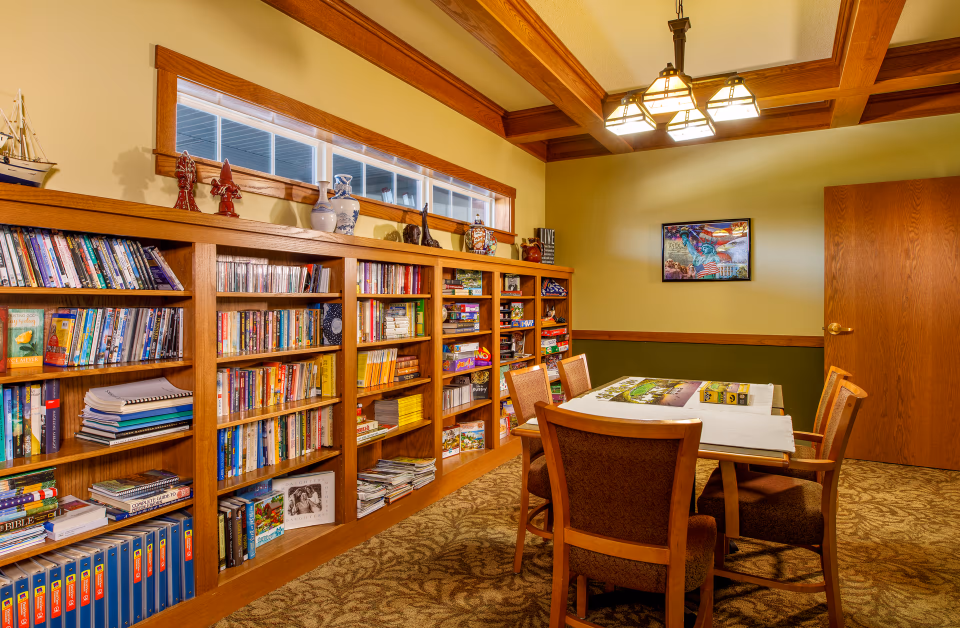 Wood-paneled community activity room with built-in bookshelves, a table and chairs set for a puzzle under a hanging light.