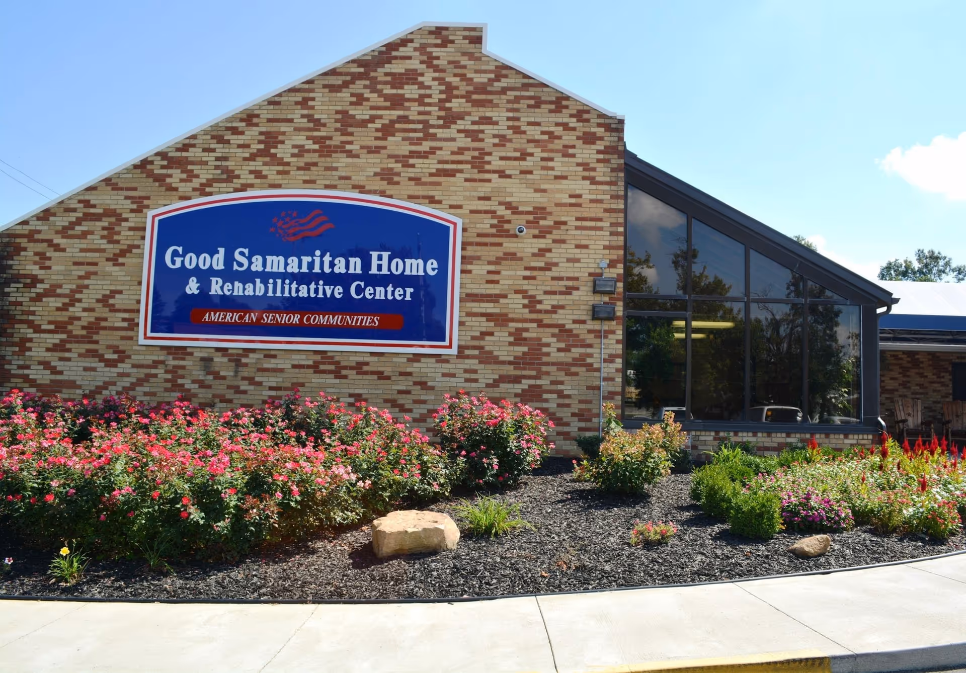 Exterior view of Good Samaritan Home & Rehabilitative Center building with a large blue and white sign on a brick wall. In front of the building, there is a landscaped garden with various flowering plants and shrubs. The sky is clear and blue.
