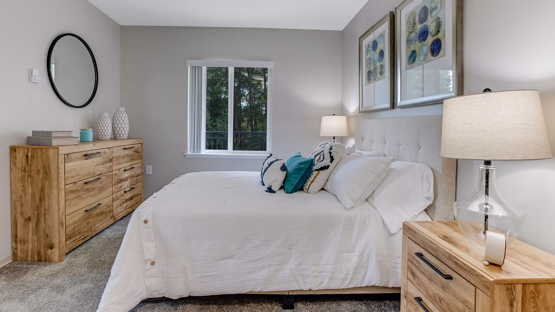 A neatly made bed with white bedding and decorative pillows in a bedroom. There is a wooden nightstand with a glass lamp on the right side of the bed. On the left side of the room, there is a wooden dresser with decorative items and a round mirror hanging above it. A window with vertical blinds shows greenery outside. Two framed abstract artworks hang above the bed.