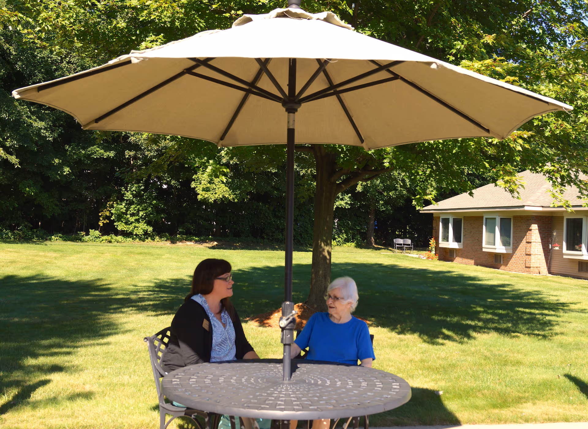 Two women sitting and talking at a round outdoor table with a large beige umbrella providing shade. They are in a grassy area with trees and a building in the background, suggesting a peaceful garden or patio setting at an assisted living center.