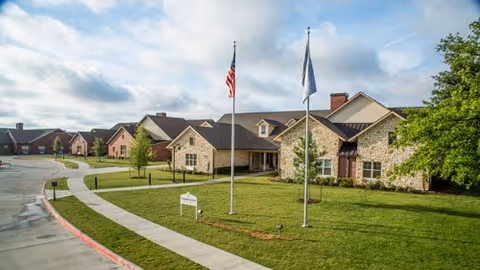 Stone-clad assisted living building with two flagpoles, a curving sidewalk and a manicured lawn under a partly cloudy sky.