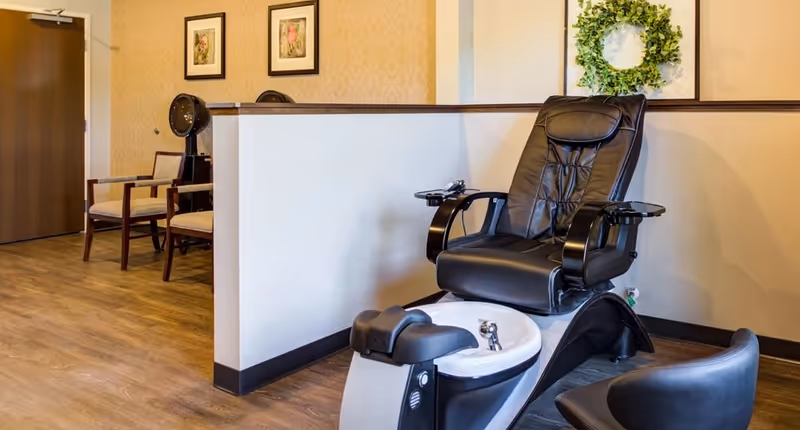Interior of a senior living facility salon area featuring a black leather pedicure chair with a foot basin, a small black chair nearby, two wooden chairs with beige cushions against a wall, a wooden door, and framed floral artwork on the wall with a decorative green wreath.