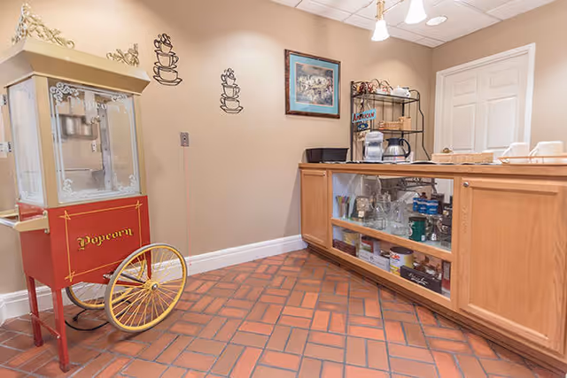 A small refreshment area with a vintage-style popcorn machine on the left and a wooden counter on the right containing coffee makers, cups, and various supplies. The floor is tiled with reddish-brown bricks, and the walls are beige with coffee cup wall decals and a framed picture.