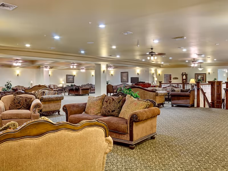 A spacious, well-lit living room area with multiple upholstered sofas and armchairs arranged for socializing. The room features patterned carpet, ceiling fans with lights, wall sconces, framed artwork, and decorative plants. There is a wooden railing and a staircase visible on the right side.