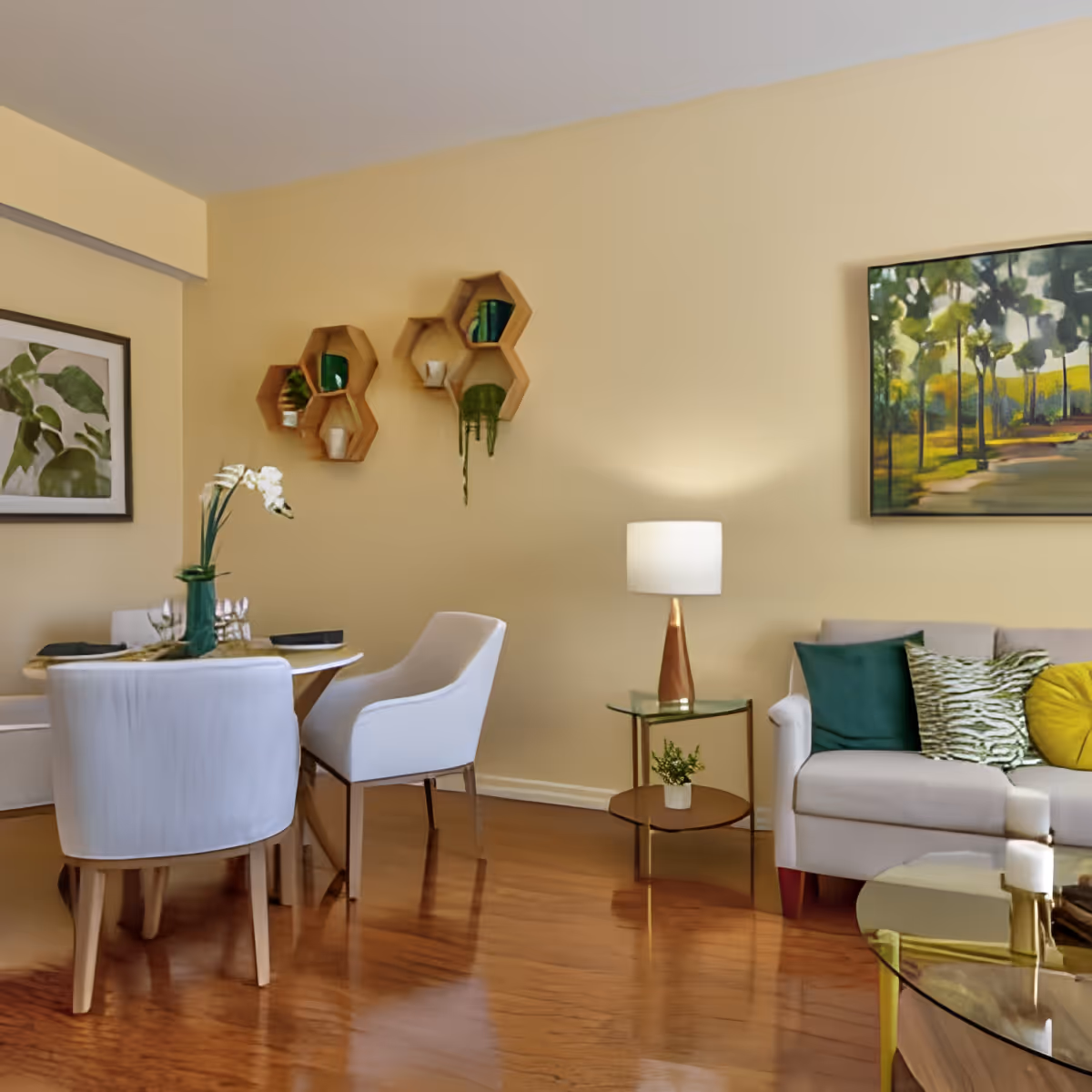 A cozy living and dining area with a small round dining table surrounded by white upholstered chairs. The walls are painted light beige and decorated with hexagonal wooden shelves holding small plants and decor items. A beige sofa with green and yellow cushions sits next to a glass side table with a lamp and a small potted plant. A framed painting of a tree-lined path hangs on the wall above the sofa. The floor is polished wood.