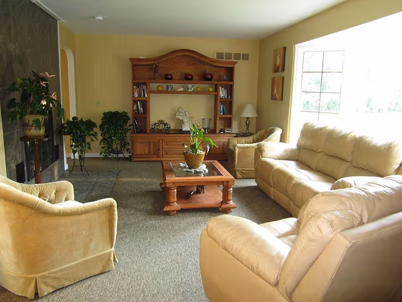 Well-lit living room with beige sofas and armchairs, a wooden coffee table and an entertainment cabinet by a large window.