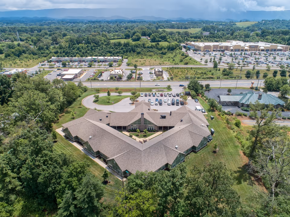 Aerial view of a senior living facility named Clover Hill Senior Living surrounded by trees and greenery. The building has a large, V-shaped roof with a parking lot in front filled with cars. In the background, there is a road, commercial buildings, and a vast landscape of fields and hills under a partly cloudy sky.