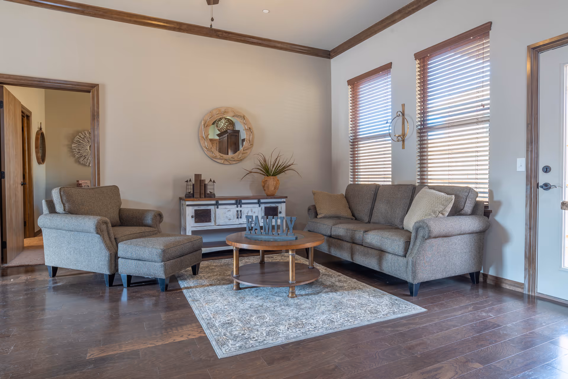 A cozy living room with a gray upholstered sofa and matching armchair with ottoman. A round wooden coffee table with a decorative 'FAMILY' sign sits on a patterned area rug. The room has wooden floors, two windows with wooden blinds, a wooden sideboard with decorative items, and a round mirror on the wall. A door with a window is visible on the right side.