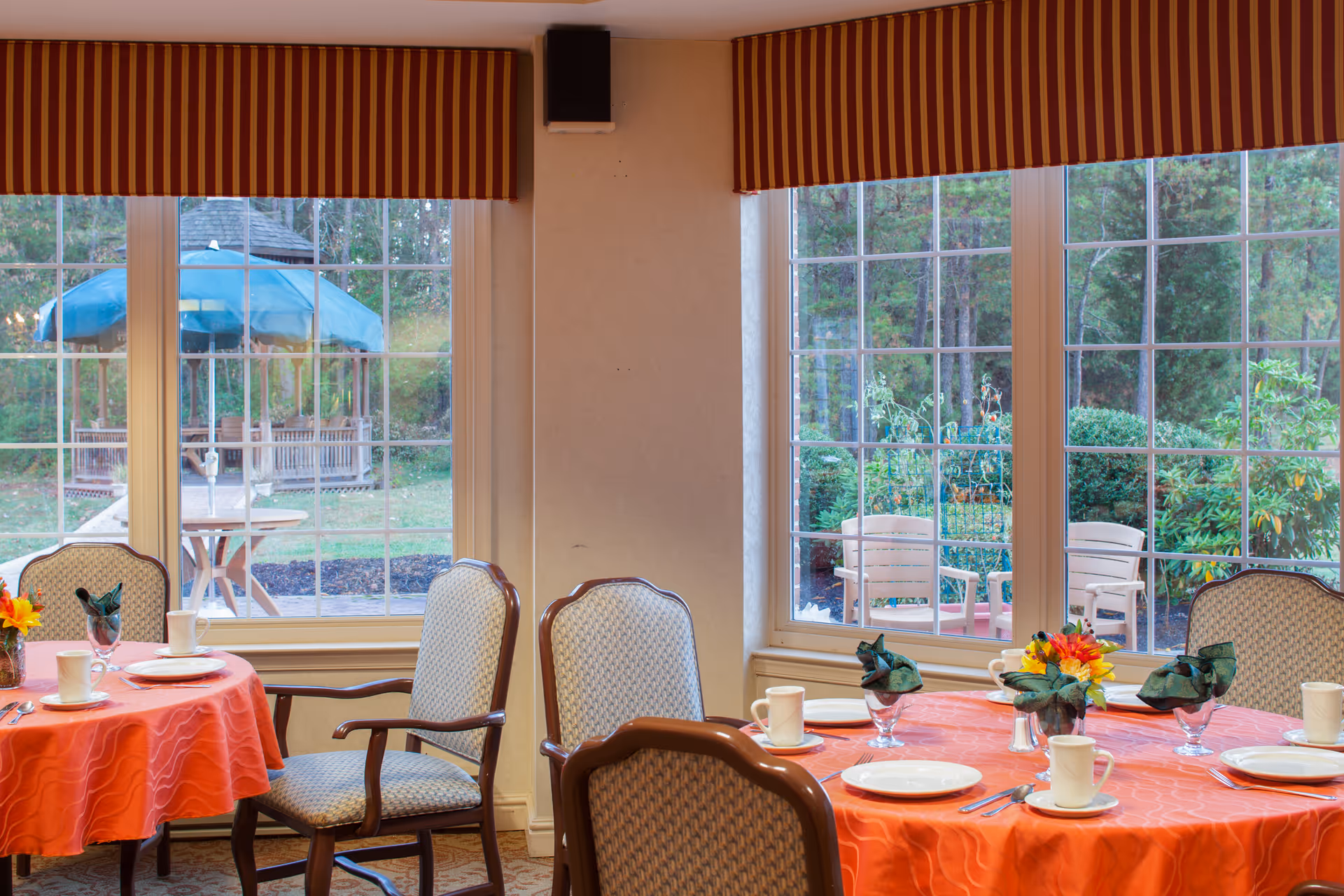 Dining area with round tables covered in orange tablecloths, set with white plates, cups, silverware, and green napkins. The room has large windows with red and yellow striped valances, showing an outdoor garden with chairs and a gazebo.