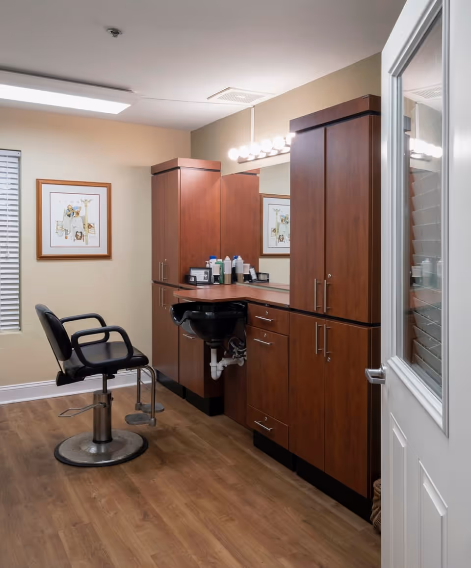 Interior view of a hair salon area in a senior living facility with a black salon chair, a black hair washing sink, wooden cabinets, a large mirror, and a framed picture on the wall. The floor is wood, and there is a door with a window partially open.
