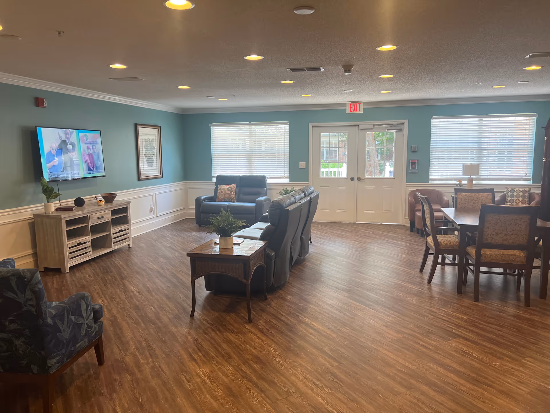 A spacious senior living community common area with wood flooring, teal walls, and white wainscoting. The room features a wall-mounted TV above a wooden media console, several comfortable chairs and sofas arranged for socializing, a wooden side table with a plant, and a dining table with chairs near windows with blinds. Double doors with glass panels lead outside.