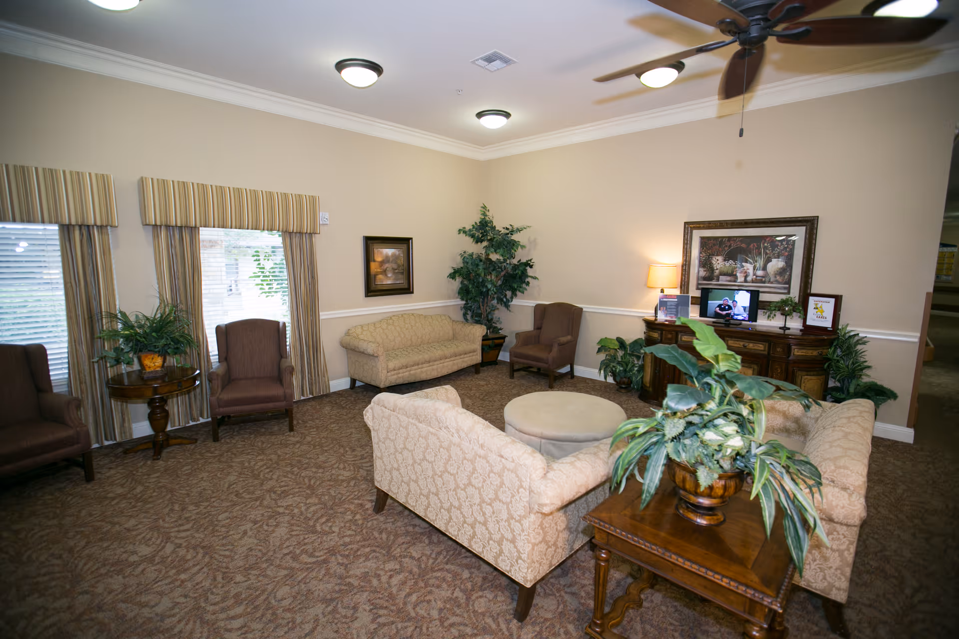 A cozy living room area in a senior living facility with beige patterned sofas, brown armchairs, a round ottoman, and a wooden side table with a plant. The room has beige walls, carpeted floor, ceiling fan, and framed artwork. A wooden cabinet with a lamp, plants, and a small TV is against one wall.