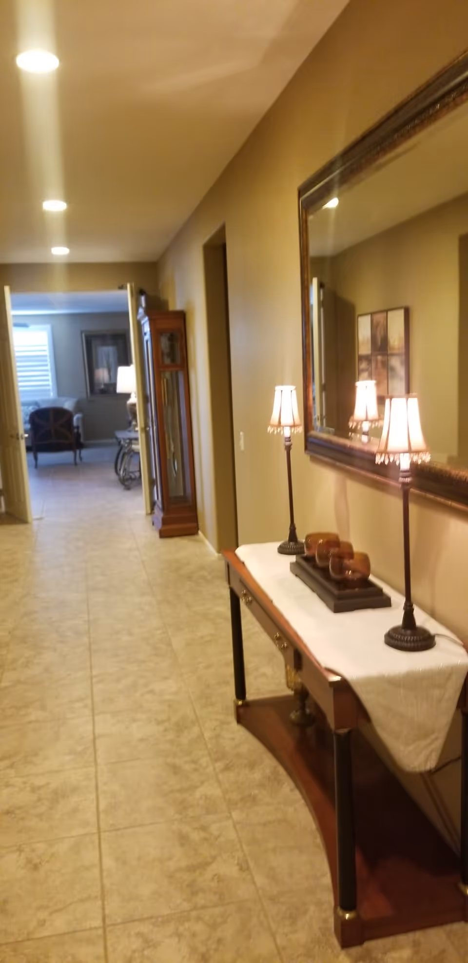 A hallway with beige walls and tiled floor leading to a living room area. On the right side of the hallway, there is a wooden console table with a white runner, two small lamps with beige shades, and decorative items. Above the table is a large framed mirror reflecting the lamps and part of the hallway. At the end of the hallway, there is a wooden grandfather clock and an open doorway leading to a living room with chairs and a window with blinds.