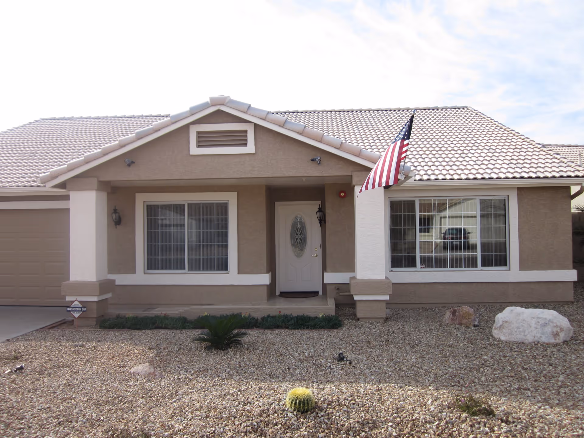 Front exterior view of a single-story house with a tiled roof, two large windows with white frames, a white front door with decorative glass, an American flag mounted on the right side of the porch, and a gravel yard with some small plants and rocks.