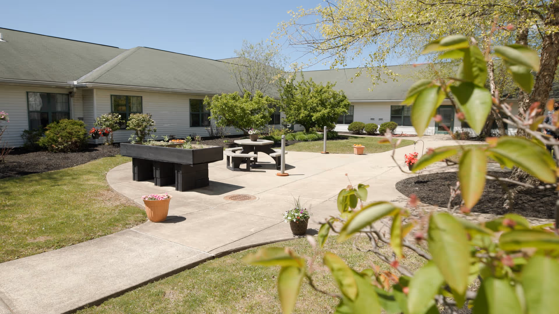Outdoor courtyard area at Stratford Commons Assisted Living featuring a concrete walkway, raised garden beds, a round picnic table with attached benches, potted flowers, and various shrubs and trees under a clear blue sky.