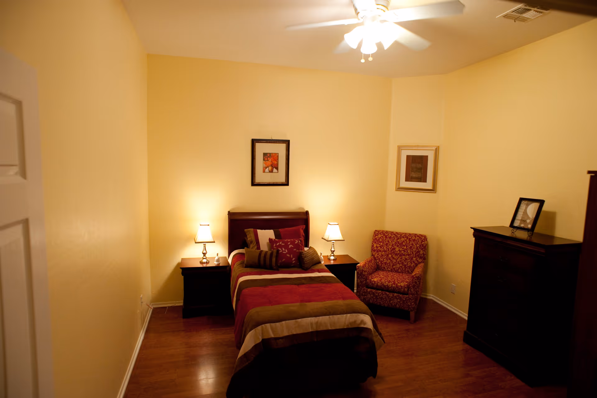 A cozy bedroom with a single bed covered in a striped red, brown, and beige comforter. There are two bedside tables with lamps on either side of the bed. A patterned armchair is placed in the corner next to a framed picture on the wall. A dark wooden dresser with a framed photo on top is on the right side of the room. The walls are painted light yellow and the floor is wooden.