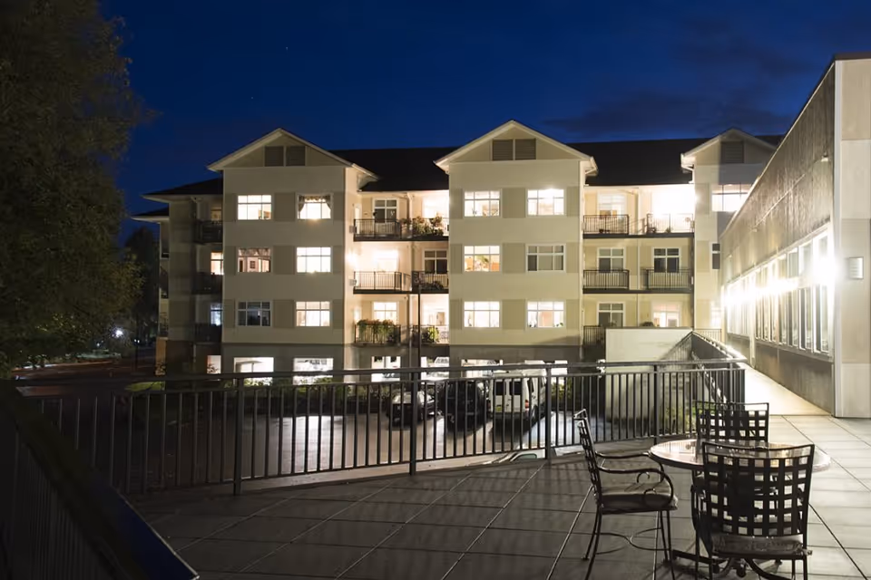 Night view of a multi-story residential building with balconies and lit windows, seen from a terrace with a round table and four metal chairs. The terrace has a railing and is adjacent to a walkway with bright wall lights.