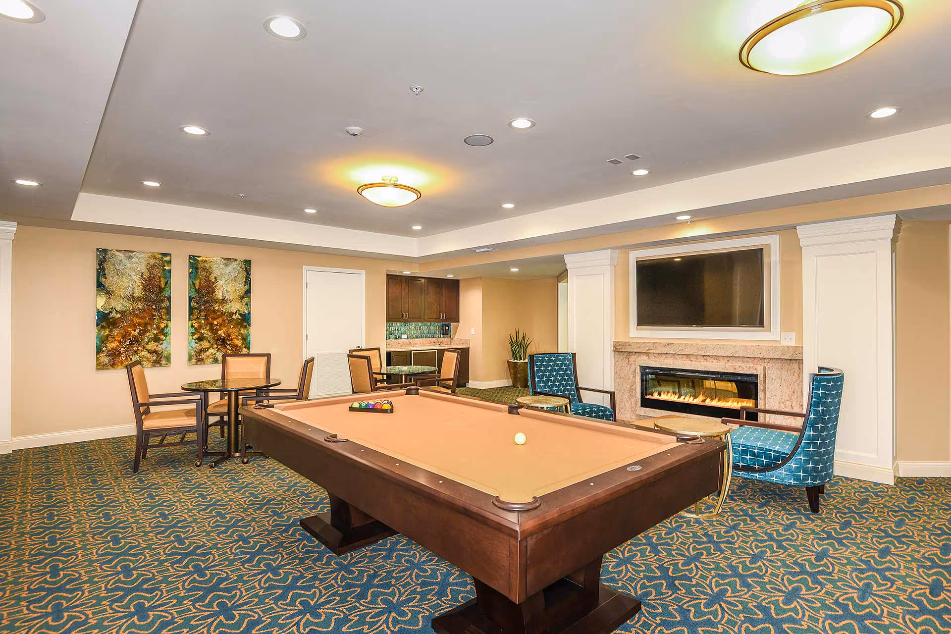 A well-lit recreational room featuring a pool table with a set of billiard balls arranged on it. The room has patterned blue carpet, beige walls, and a ceiling with recessed lighting and a central light fixture. There are two blue patterned armchairs and a fireplace with a mounted flat-screen TV above it. In the background, there are two small round tables with chairs and a small kitchenette area with cabinets and a countertop.