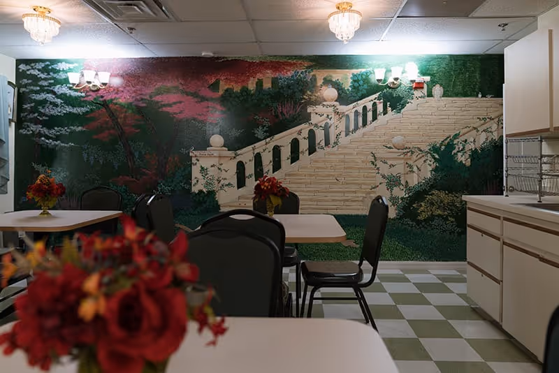 Interior view of a dining area in an assisted living facility with tables and chairs. The back wall features a large mural depicting a garden scene with stairs and greenery. There are floral centerpieces on the tables and ceiling lights providing illumination.