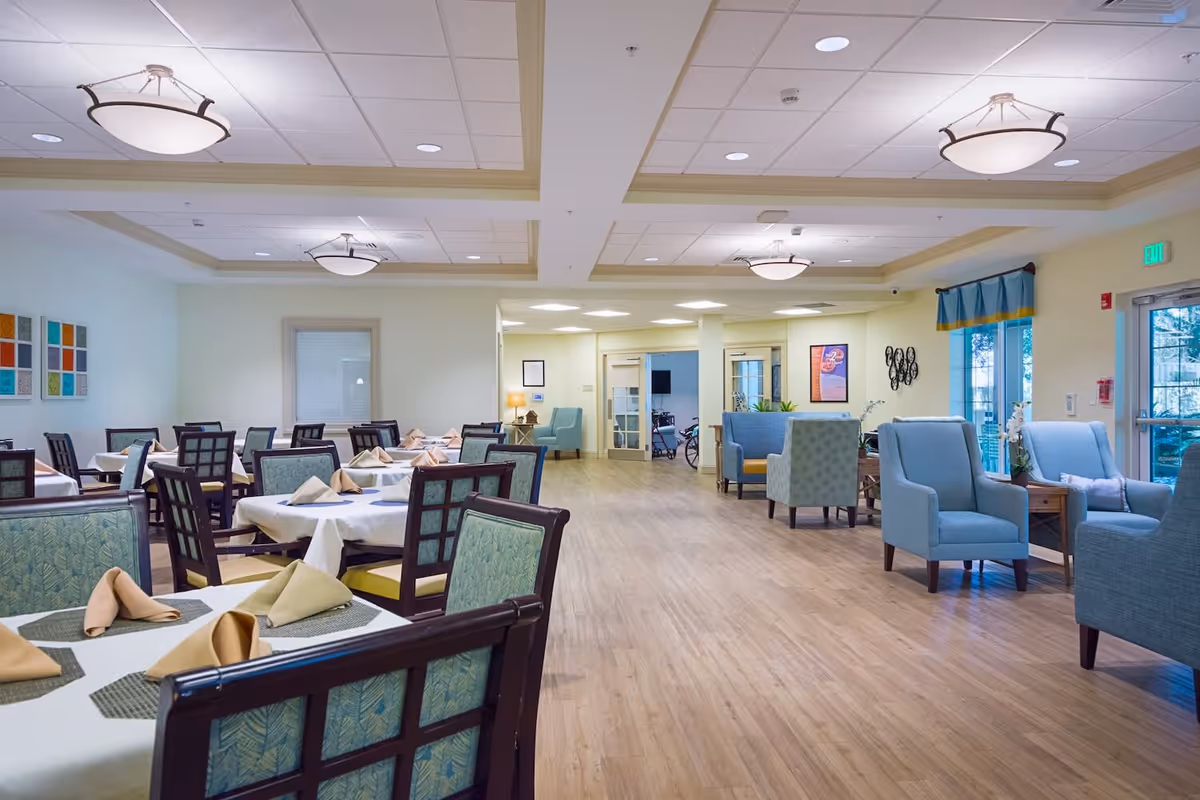A spacious dining area with multiple tables covered in white tablecloths and beige napkins, surrounded by cushioned chairs with patterned upholstery. To the right, there is a seating area with blue armchairs and small wooden tables. The room has a light wood floor, soft yellow walls, and ceiling lights. Large windows with blue curtains allow natural light to enter.