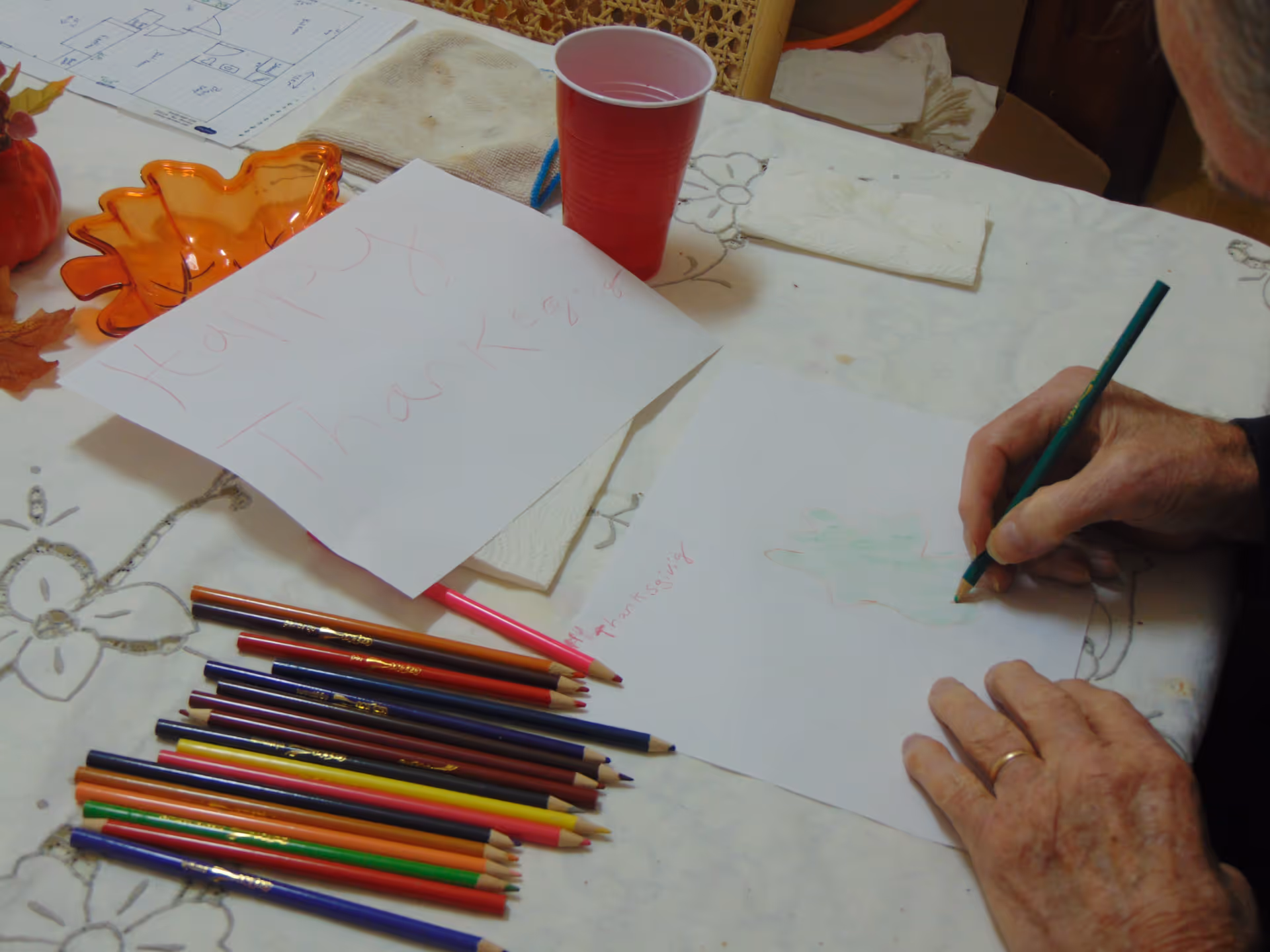 An elderly person is coloring a leaf shape on a piece of paper using a green colored pencil at a table. The table is covered with a white tablecloth with floral patterns. There are multiple colored pencils laid out on the table, a red plastic cup, a small orange leaf-shaped dish, and a paper with 'Happy Thanksgiving' written on it.