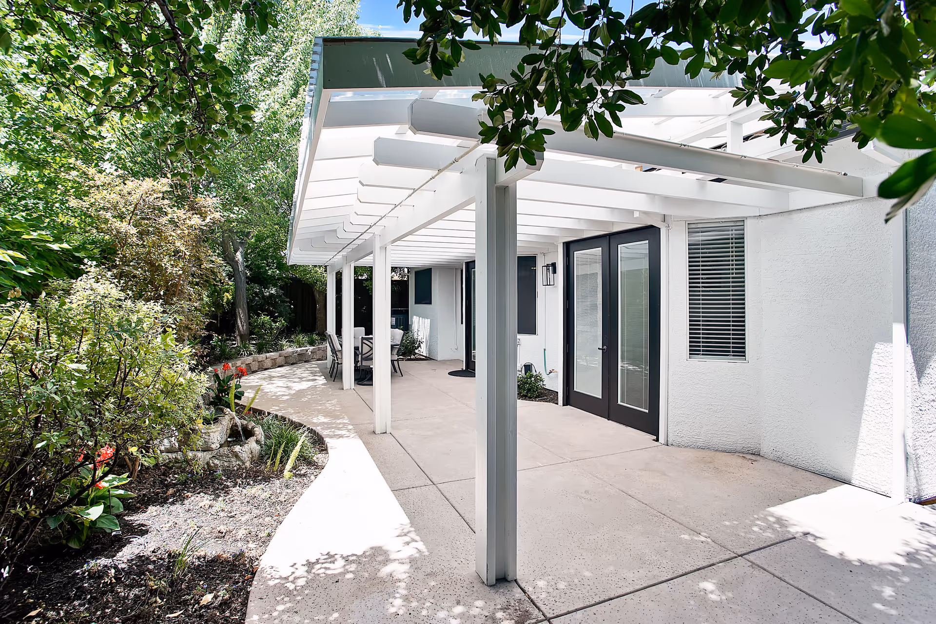 Outdoor patio area with a white pergola attached to a building. The patio has a concrete floor and is surrounded by lush green trees and plants. There is a table with chairs under the pergola near the building's glass doors and windows.