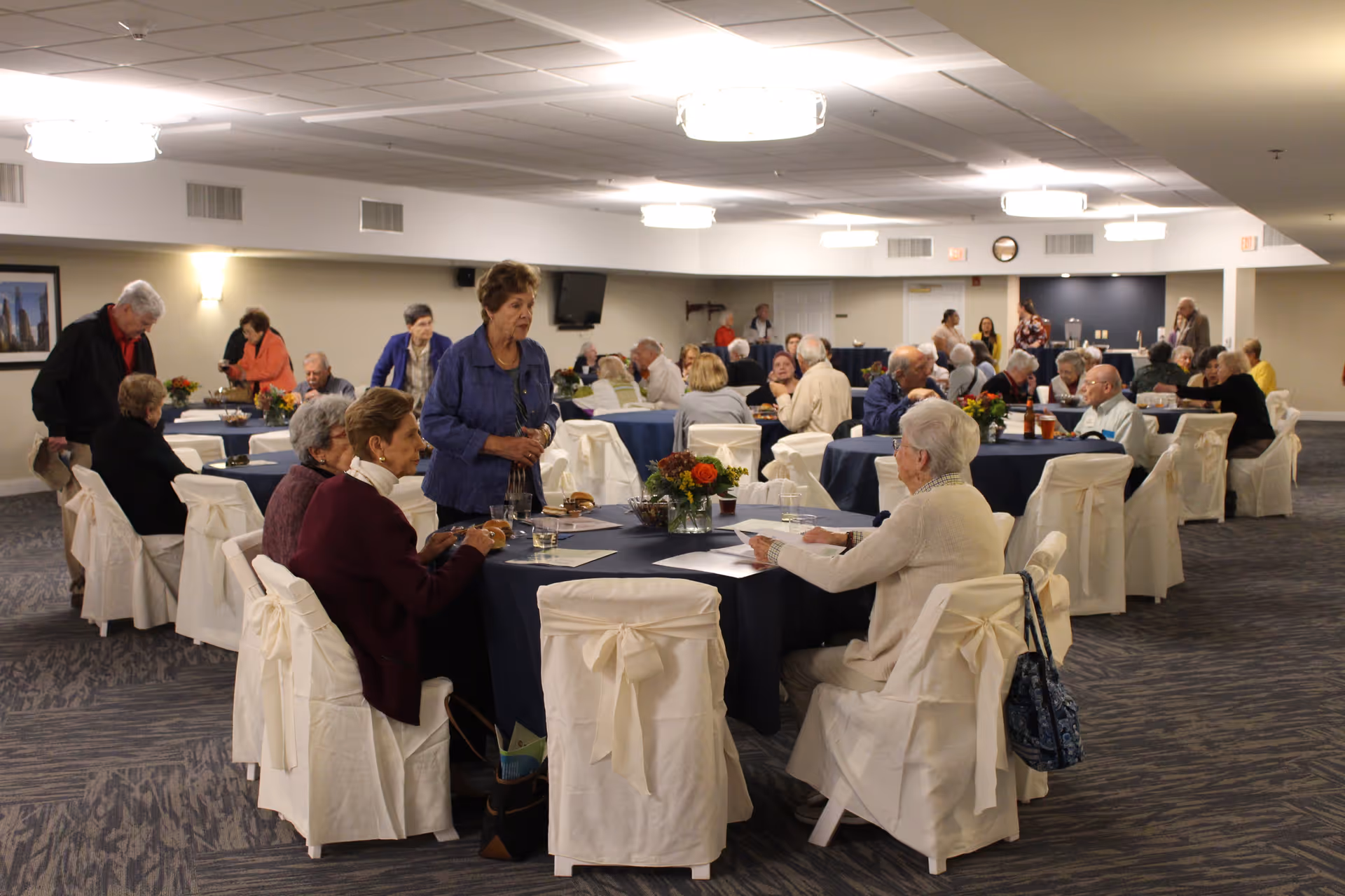 A large room with multiple round tables covered with dark blue tablecloths and white chairs with white covers and bows. Several elderly people are seated and standing around the tables, engaging in conversation and socializing. The room has a carpeted floor, white ceiling with recessed lighting, and neutral-colored walls. There are flower centerpieces on the tables and a few people in the background near a counter area.