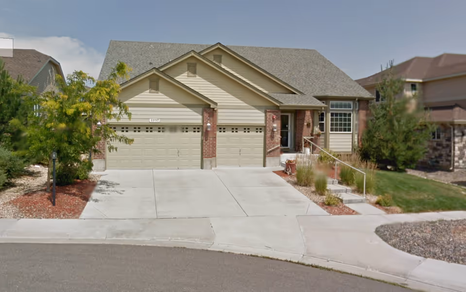 Front exterior view of a single-story residential building with three garage doors, a small staircase leading to the entrance, and landscaping with trees and shrubs.
