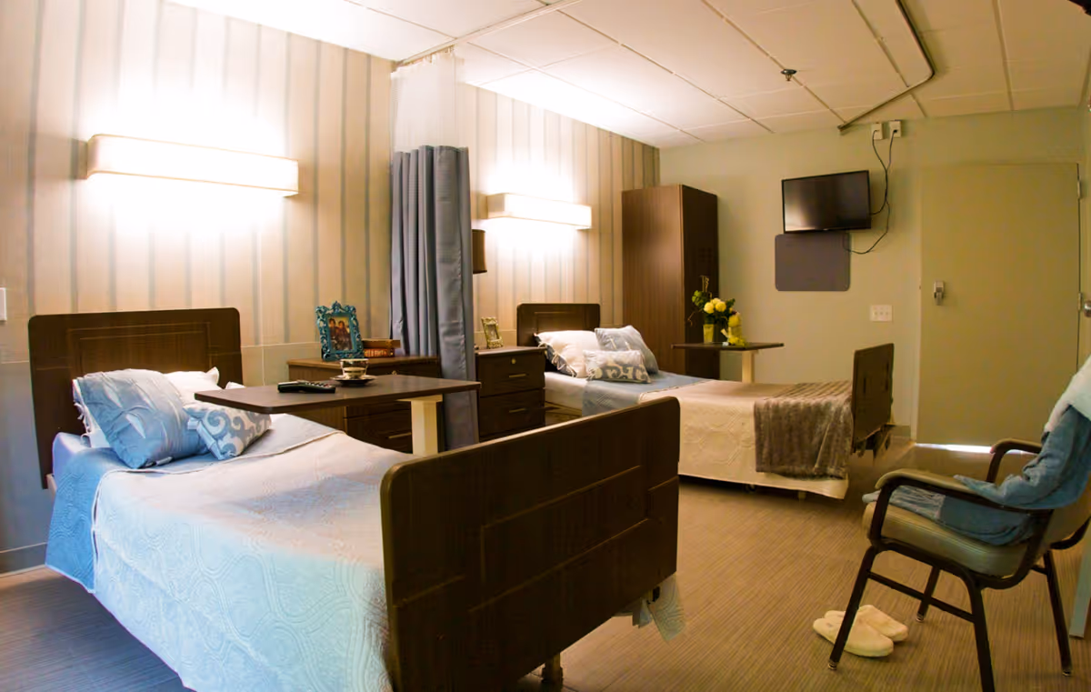 A nursing center room with two hospital-style beds, each with pillows and blankets. There is a wooden nightstand between the beds with framed photos and a remote control. A curtain divider is partially drawn between the beds. A TV is mounted on the wall opposite the beds, and a cushioned chair with a blanket is positioned near the door. The room has soft lighting and a calm, neutral color scheme.