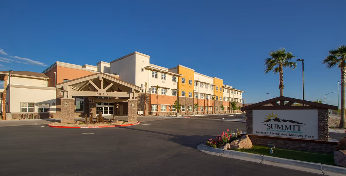 Exterior view of a multi-story senior living facility building with a covered entrance and a sign in front that reads 'The Summit Assisted Living and Memory Care'. The building has a mix of beige, white, and yellow colors with several windows, palm trees, and a clear blue sky.