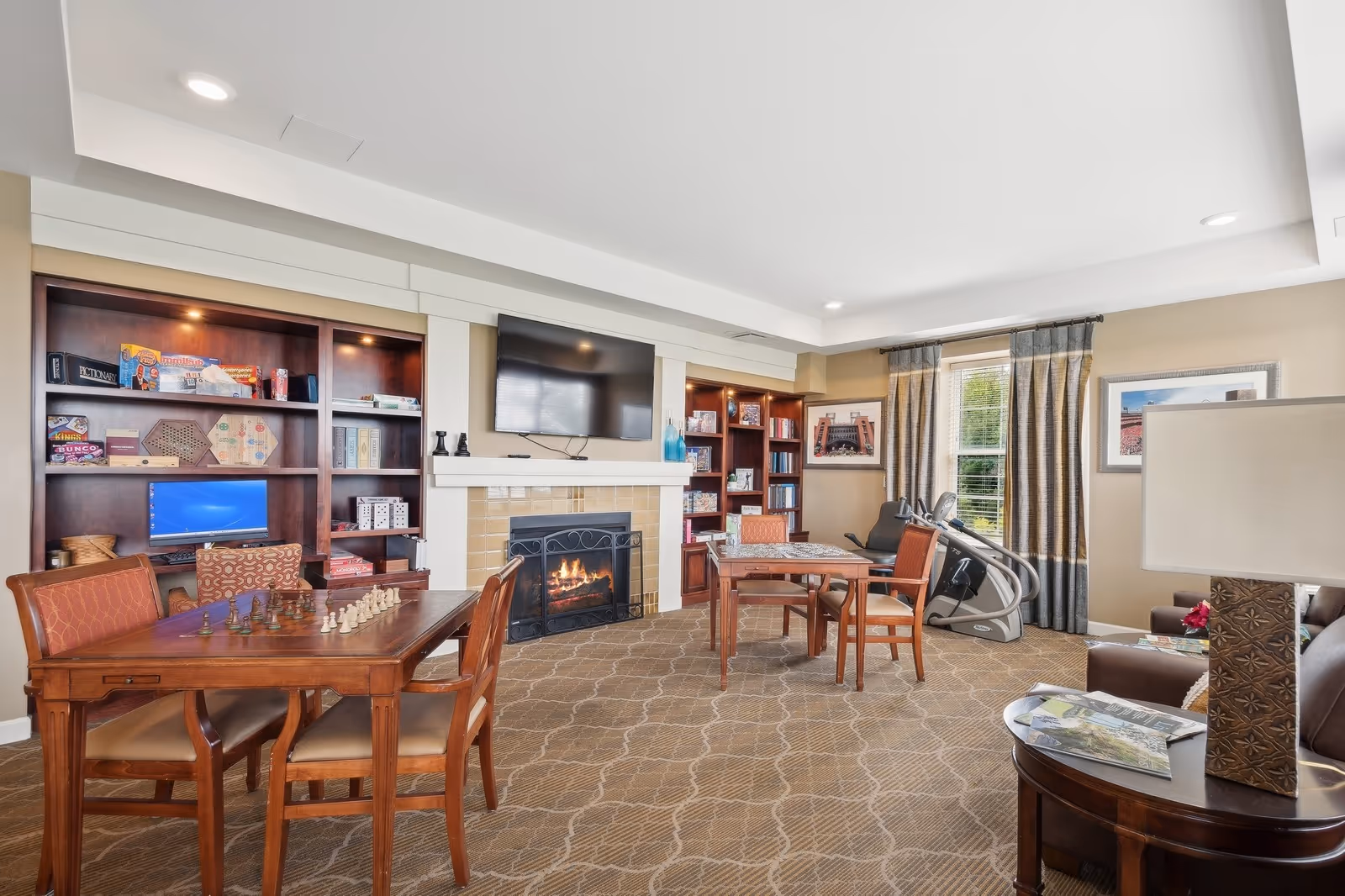 A cozy common room with a fireplace and a mounted TV above it. There are two wooden tables with chairs around them, one table has a chess set and the other has a puzzle. Built-in wooden shelves filled with board games, books, and a computer monitor flank the fireplace. A window with curtains lets in natural light, and an exercise machine is positioned near the window. A side table with a lamp and magazines is visible in the foreground.