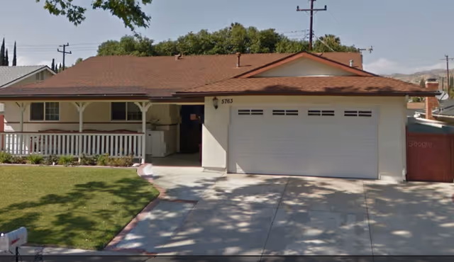 Front exterior view of a single-story residential building with a brown shingle roof, white walls, a two-car garage, a covered porch with white railing, and a driveway leading up to the garage. The house number 5763 is visible above the garage door. There is a green lawn and some shrubs in front of the porch.