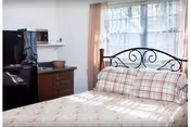 A small bedroom with a bed featuring a metal headboard and plaid bedding. Next to the bed is a kitchenette area with a black refrigerator, microwave, and wooden cabinets. A window with sheer curtains allows natural light to fill the room.