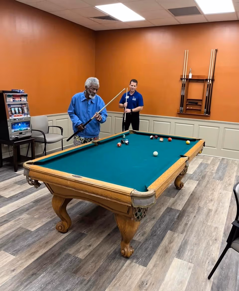 Two men playing pool in a recreation room with orange walls and wood paneling. One man is preparing to take a shot while the other watches. There is a slot machine and chairs in the background, and a rack with pool cues mounted on the wall.