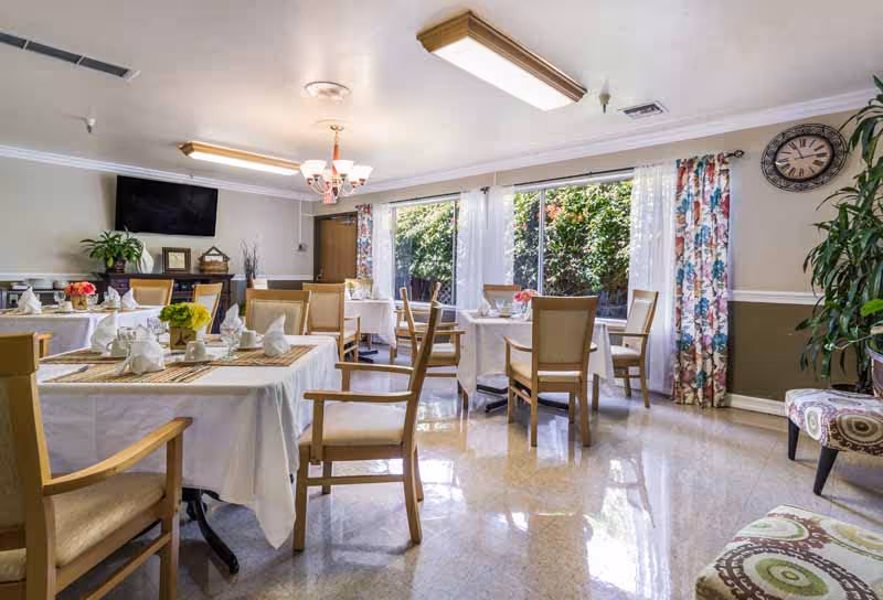 A bright dining room with several tables covered in white tablecloths, each set with napkins and small floral centerpieces. Wooden chairs surround the tables. Large windows with floral curtains let in natural light, and a wall clock and potted plants decorate the room.