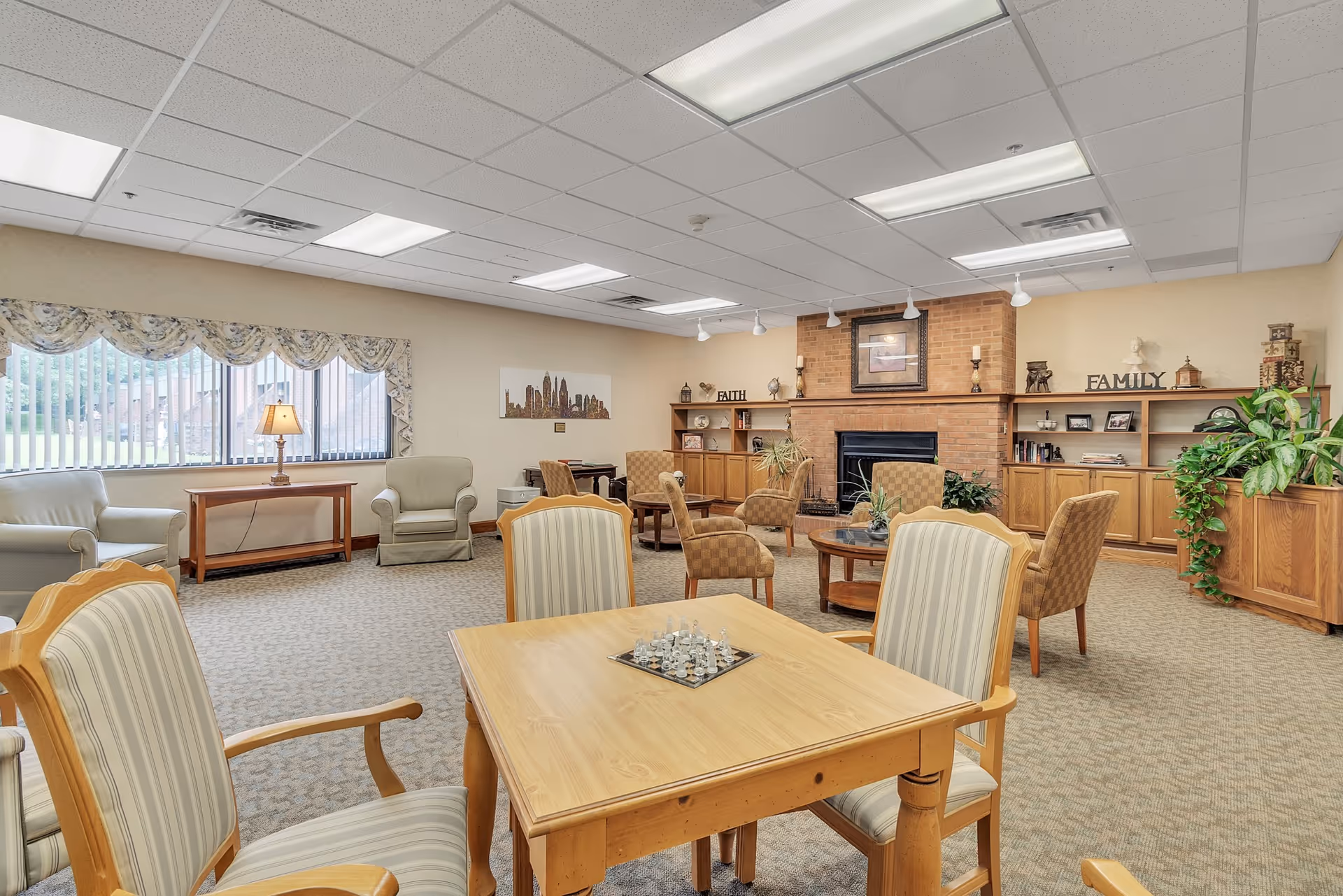 A spacious living room area in a senior living facility with a wooden table and four striped cushioned chairs in the foreground, a chess set on the table, several armchairs and sofas arranged around the room, a brick fireplace with shelves on either side decorated with plants, books, and decorative items, and large windows with vertical blinds letting in natural light.